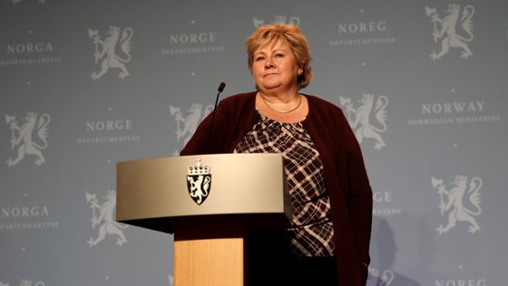 Statsminister Erna Solberg står ved talerstolen på pressekonferansen, foran blå vegg med riksløver og departementslogoer.