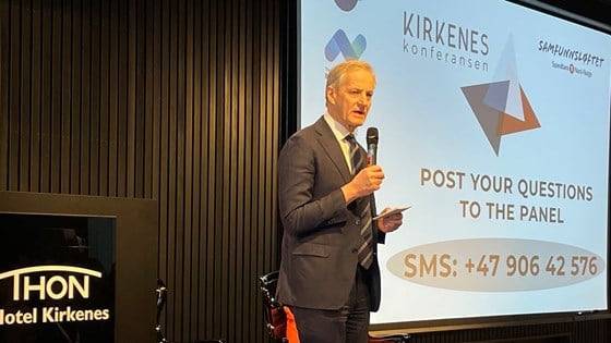Statsministeren står på en scene og foran et lerret med Kirkenes-konferansen-logo. Han er kledd i dress og holder en mikrofon.