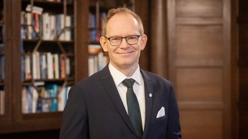 Portrett av en smilende forsknings- og høyere utdanningsminister Oddmund Hoel, som står i dress foran en brun bokhylle.