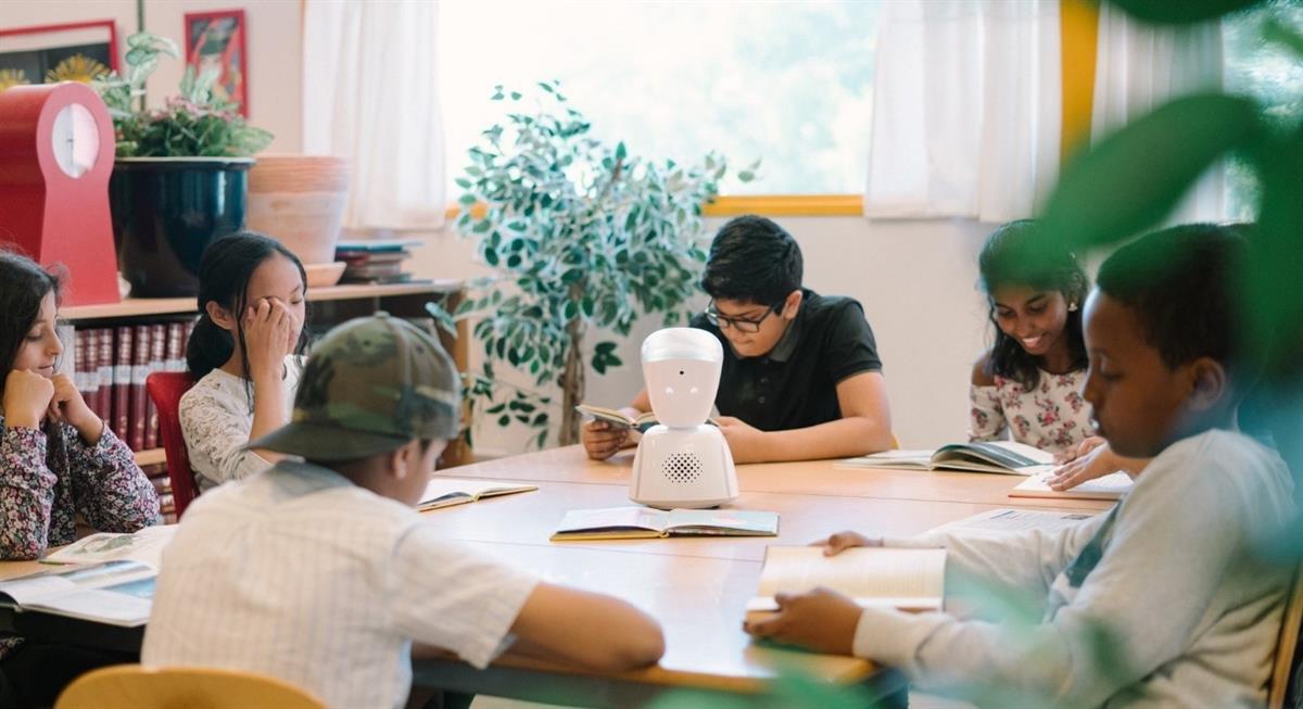 Six children sitting around a table doing school work. The robot AV1 sits in the centre of the table.