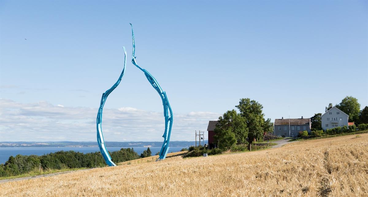 Landscape from the Østre Toten region. A farm and fields by the lake Mjøsa in the background and a sculpture in the foreground.
