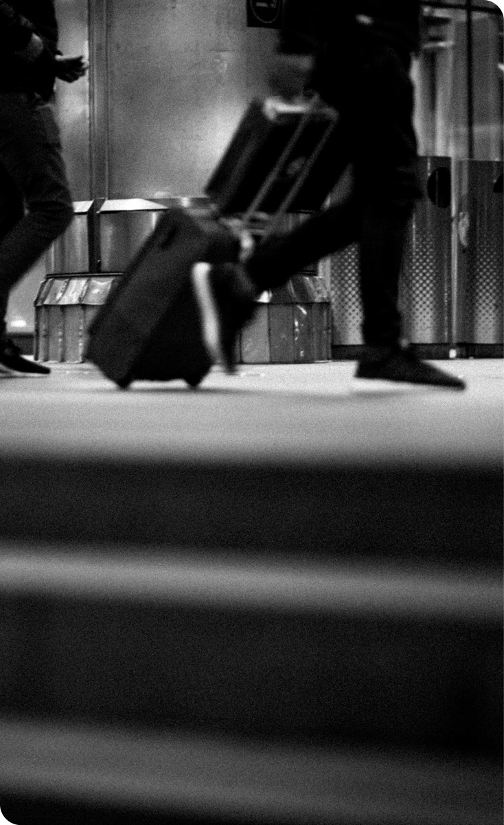 Svart/hvitt-bilde fra et trafikknutepunkt. Muligens en togstasjon. Bildet viser utsnitt av mennesker hvor du ser bena til folk som går. Han som er mest framtredende, har en trillekoffert etter seg.