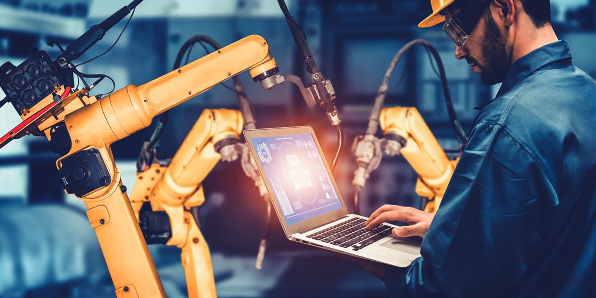 Bilde av en mann med hjelm og vernebriller som arbeider på en datamaskin i et fabrikklokale.