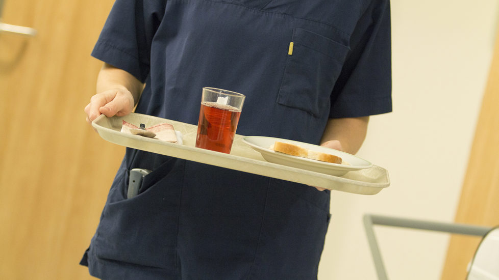 Vi ser overkroppen til en helsearbeider, men ikke hodet. Vedkommende er ikledd uniform og holder et brett med et glass saft.