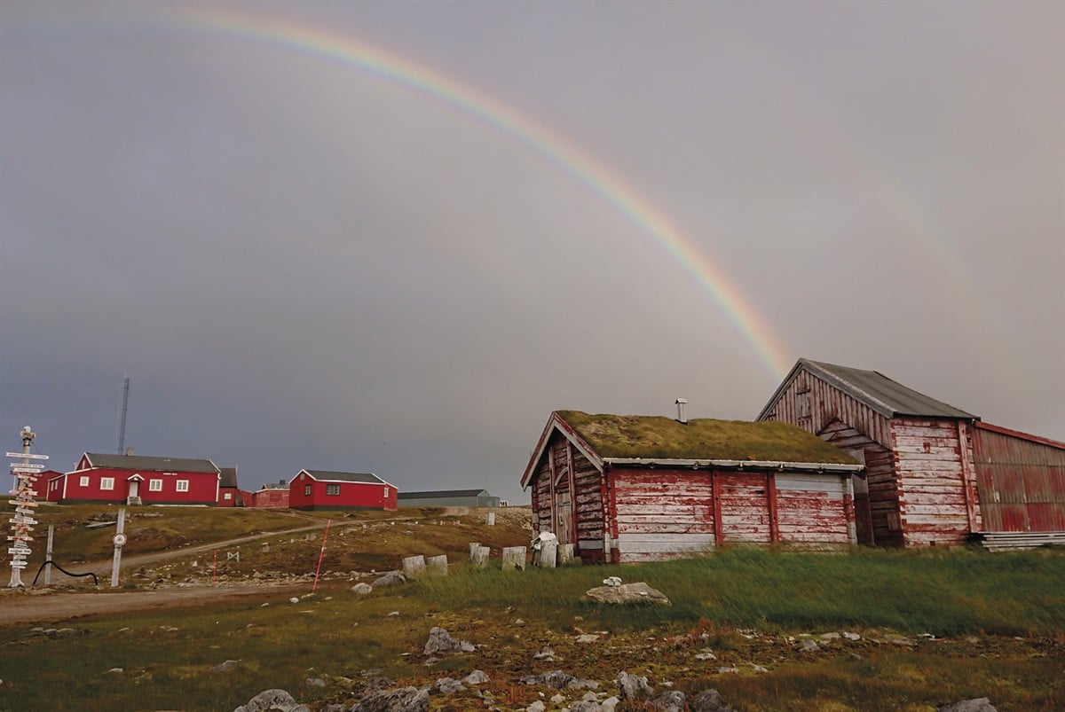 Røde bygninger på høyfjellet, der den nærmeste har torvtak. Det er overskyet, og en regnbue er synlig på himmelen.