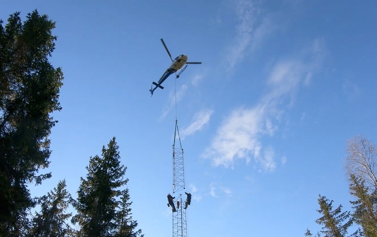To som klatrer i en telemast som monteres. Et helikopter flyr over med en del som skal monteres.