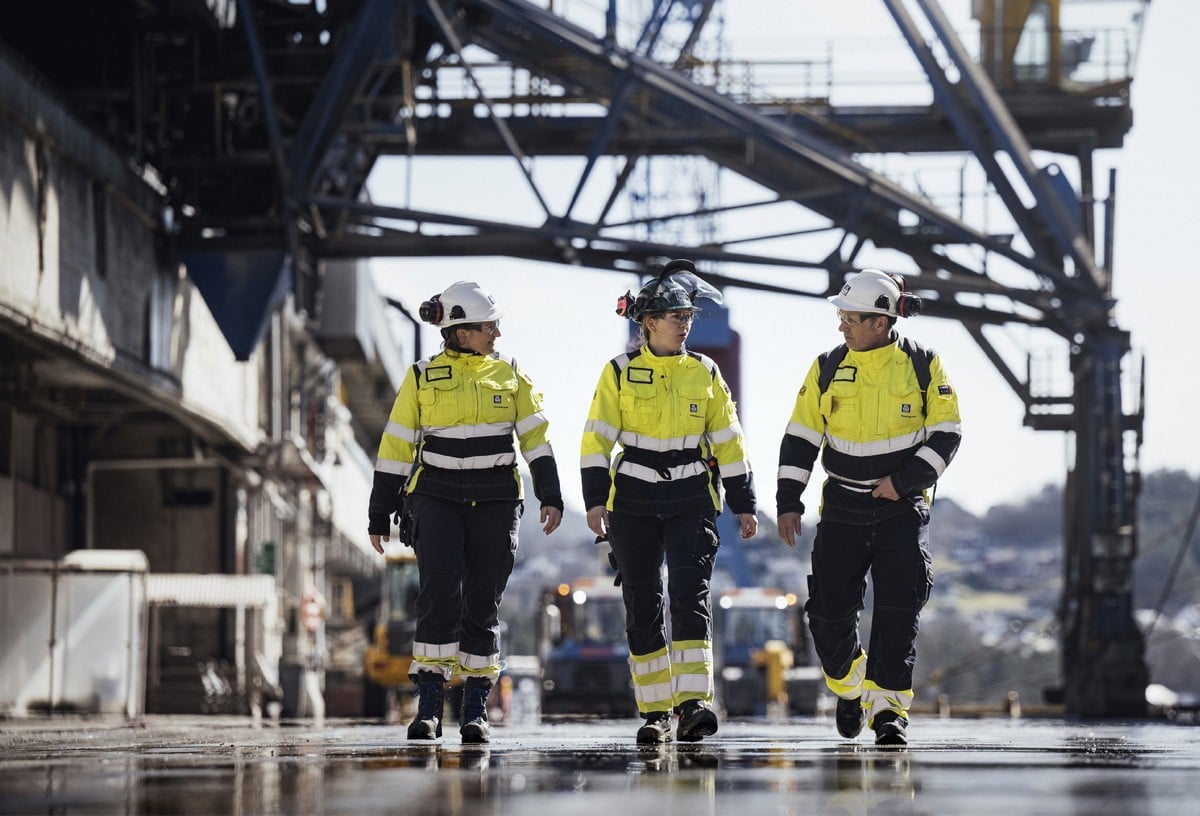 Tre arbeidere i arbeidsklær som går inne på et industriområde.