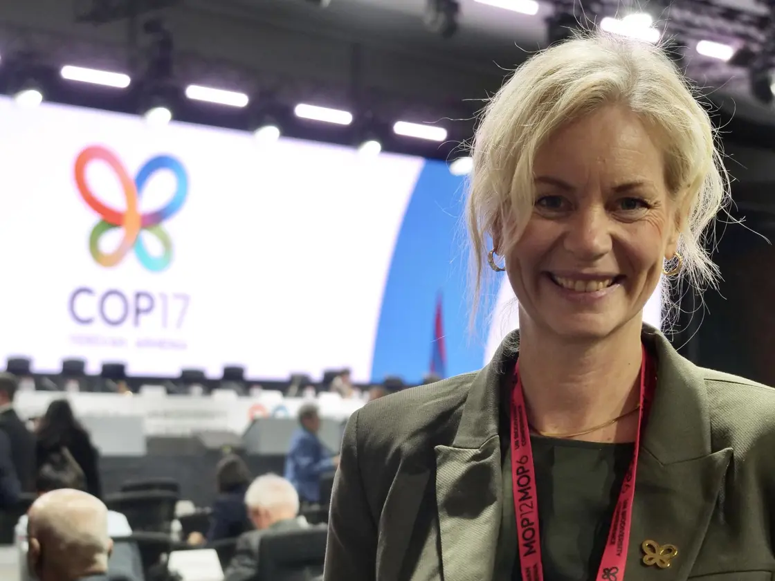 Bilde av Sigrun Aasland som smiler mot kamera. I bakgrunnen ein enorm skjerm på ei scene der det står COP17.