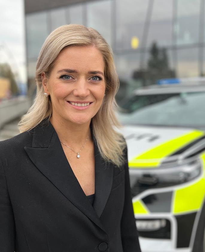 En smilende justis- og beredskapsminister Emilie Enger Mehl, iført sort jakke, ser inn i kamera. En politibil  og et bygg med glassfasade skimtes i bakgrunnen.
