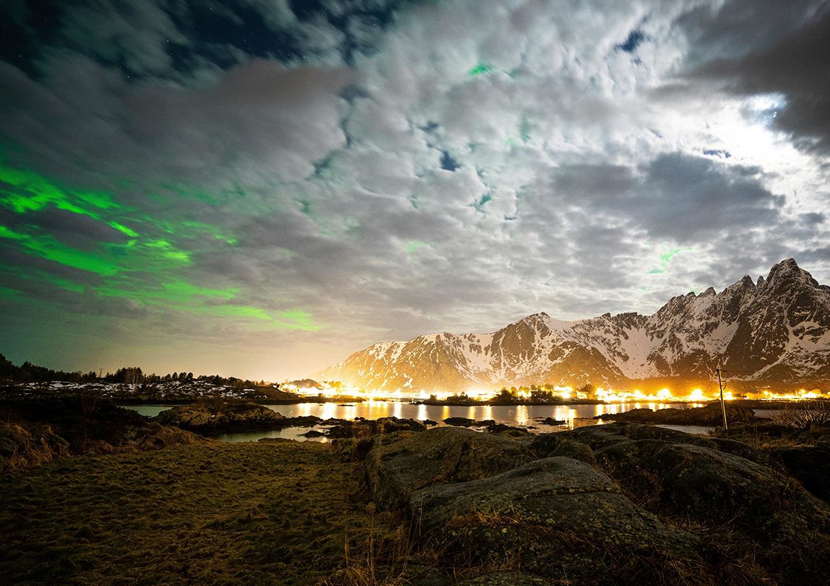 Naturbilde tatt på nattestid med sterk belysning fra begynnelse som ligger langs kystlinje og en himmel som også lyses opp av Nordlys.