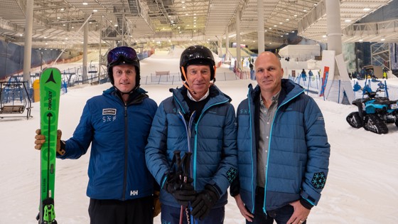 På bildet ser du tre menn i vintertøy foran en skibakke. Skibakken er innendørs.