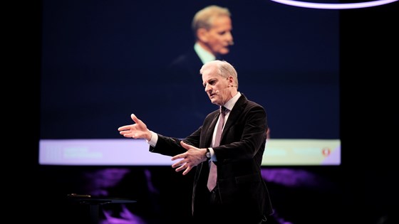 Statsminister Jonas Gahr Støre står på scenen, gestikulerer. Kledd i dress med slips. I bakgrunnen ser man Støre fra en annen vinkel på en storskjerm.