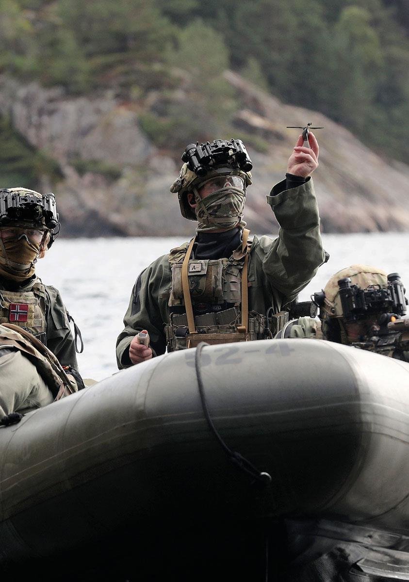 A masked soldier sits in an inflatable boat on the fjord, facing the camera. His arm is raised. In his hand is a micro uncrewed aerial vehicle (micro-UAV) that is about to take off.