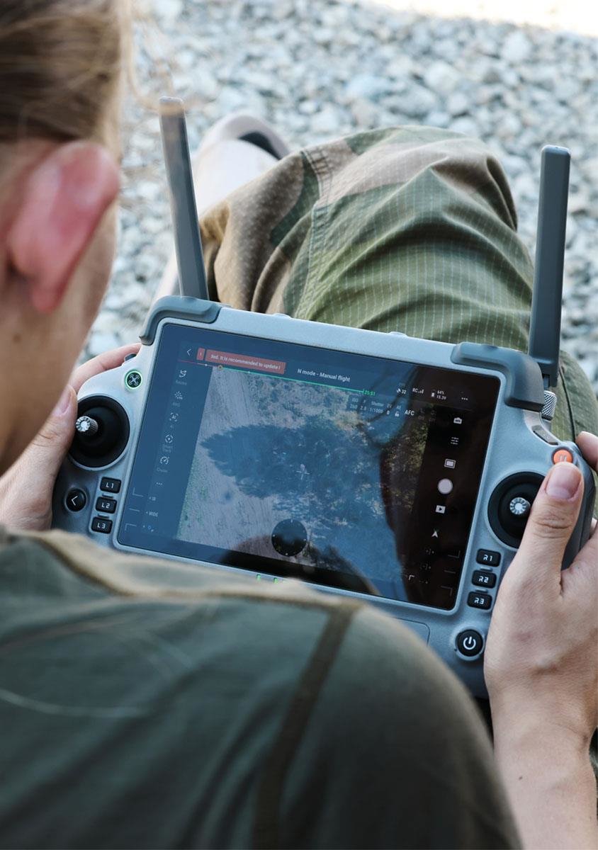 A glance over the shoulder of a soldier concentrating on operating the remote control of an uncrewed aerial system (UAS).