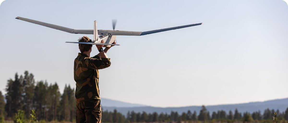 En soldat står på en åsrygg med ryggen mot kameraet. I hendene holder soldaten en drone over hodet som ligner et modellfly, klar for å hjelpe det til å lette.