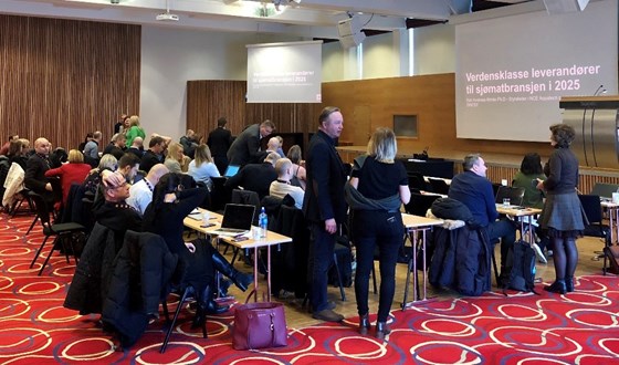 Bildet viser en møtesal med mange deltakere.