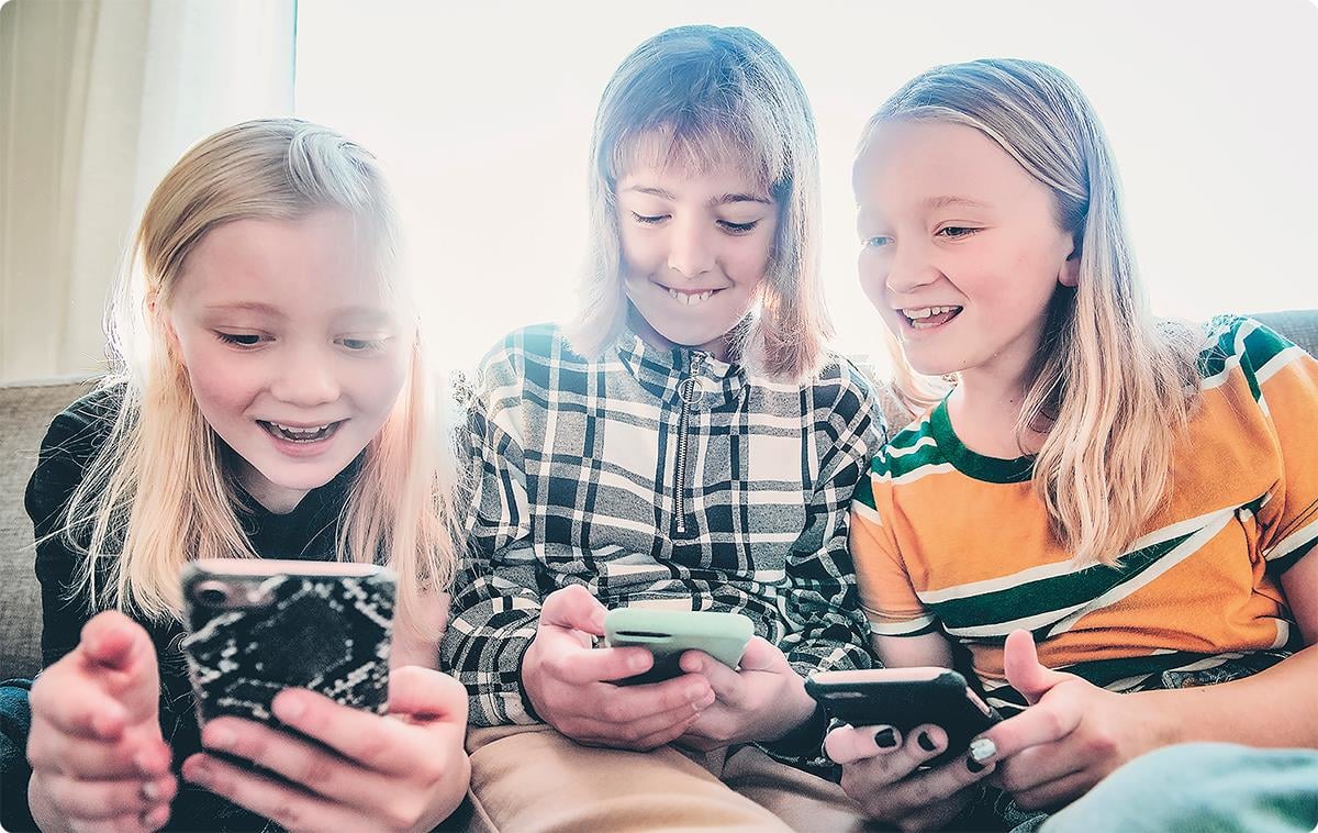 Jenter på en sofa som ser på mobiltelefoner og smiler.