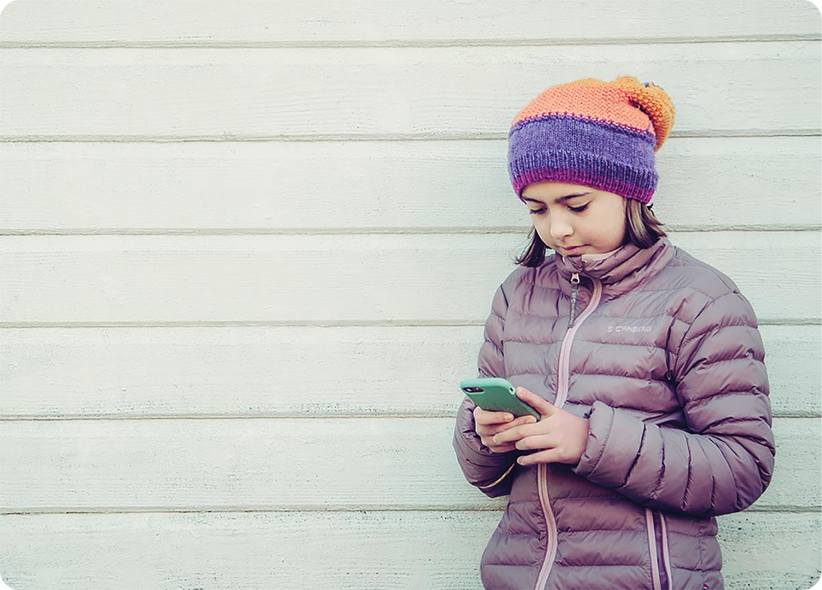 Jente med vinterjakke og lue, lent mot en vegg, som ser på mobiltelefon.