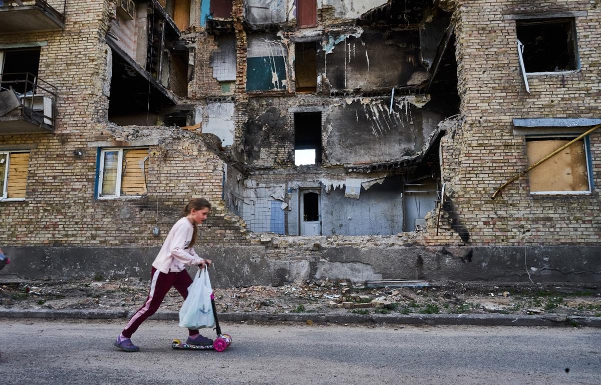 En jente sparkesykler forbi en ødelagt blokk i landsbyen Horenka i Ukraina. På styret henger en handlepose. Foto: Iva Zimova/Panos Pictures/Felix Features