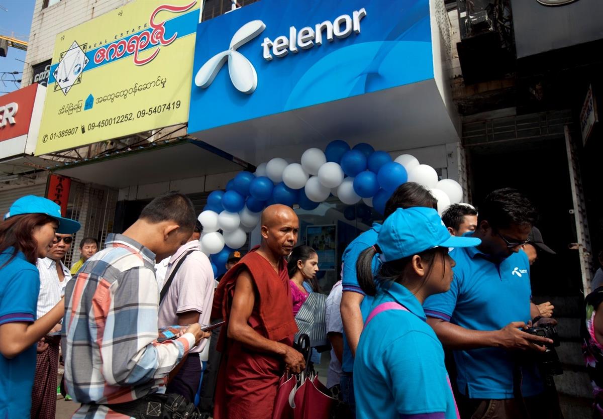 En buddistmunk står i kø sammen med andre kunder foran en butikk i 2014. Telenors logo er synlig over hodene deres, inngangen er prydet av hvite og blå ballonger i en bue. Foto: Khin Maung Win/AP/NTB