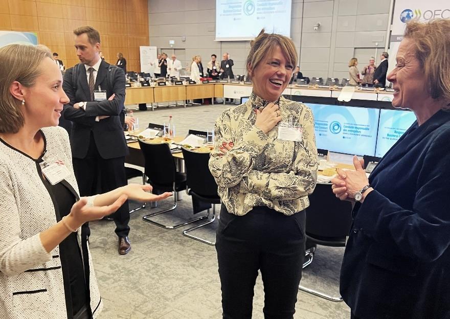 Tre deltakere i samtale, i en pause i OECDs ministermøte. I bakgrunnen ser vi pulter og stoler plassert i en massiv U-form i det store rommet. Foto: Margrethe Rosbach/NFD