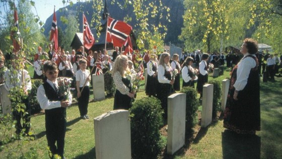 Barn i bunad og med norske flagg stilt opp ved graver på 17. mai-markering ved krigsgraver på Nes kirkegård.
