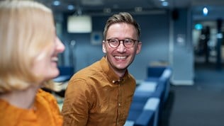 Ung smilende mann med briller i midten av bilde vendt mot ung smilende kvinne som sees uskarpt i forgrunnen til venstre i bildet. De er i et kontormiljø.