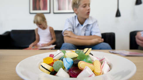 Et bilde av en godteriskål på et bord i forgrunnen, en gutt og en jente sitter på en sofa i bakgrunnen