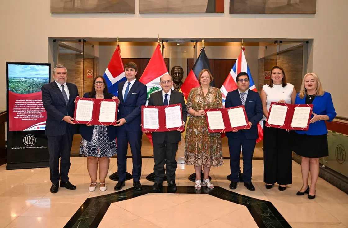 Åtte personer står og holder signerte kontrakter. I bakgrunn er det norsk, peruviansk, tysk og britisk flagg.