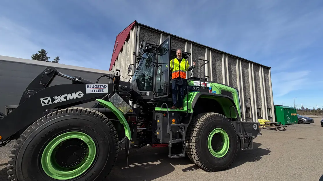 Statsråden med refleksvest som svinger seg i en stor hjullaster på et grusområde. Maskinen er grønn med store dekk og førerhuset er synlig fra siden. I bakgrunnen er det åpent område og trær