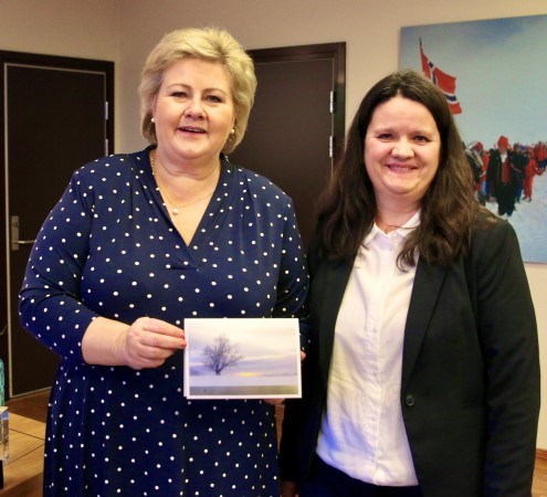 Statsminister Erna Solberg og Cathrine Strømø står ved siden av hverandre på statsministerens kontor, og viser fram årets julekort fra statsministeren.