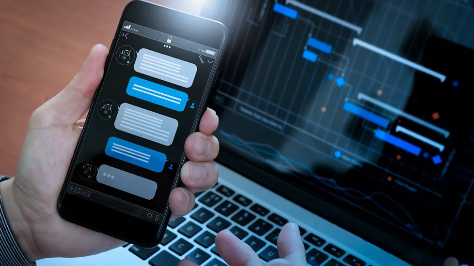 En hånd holder en mobiltelefon med en chattedialog på skjermen - en laptop er i bakgrunnen