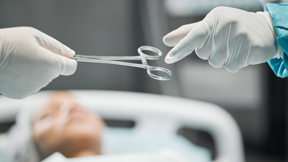 Vi ser en hånd med kirurgisk hanske på gi en saks til en annen kirurg med hanske på. I bakgrunnen ligger en pasient med gjenlukkede øyne.