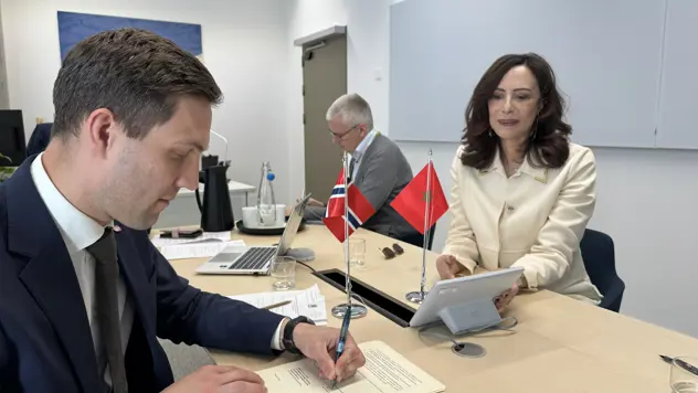 Tre personer sitter rundt et bord med det norske og marokkanske flagget. Et dokument blir signert.