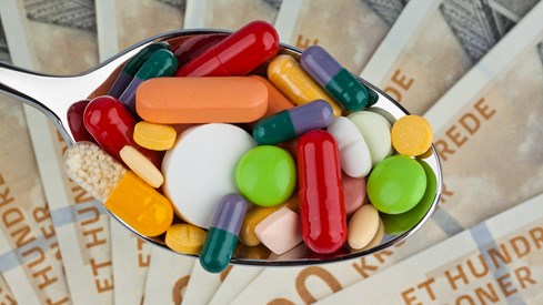 En skje med piller i ulike farger med pengesedler i bakgrunnen