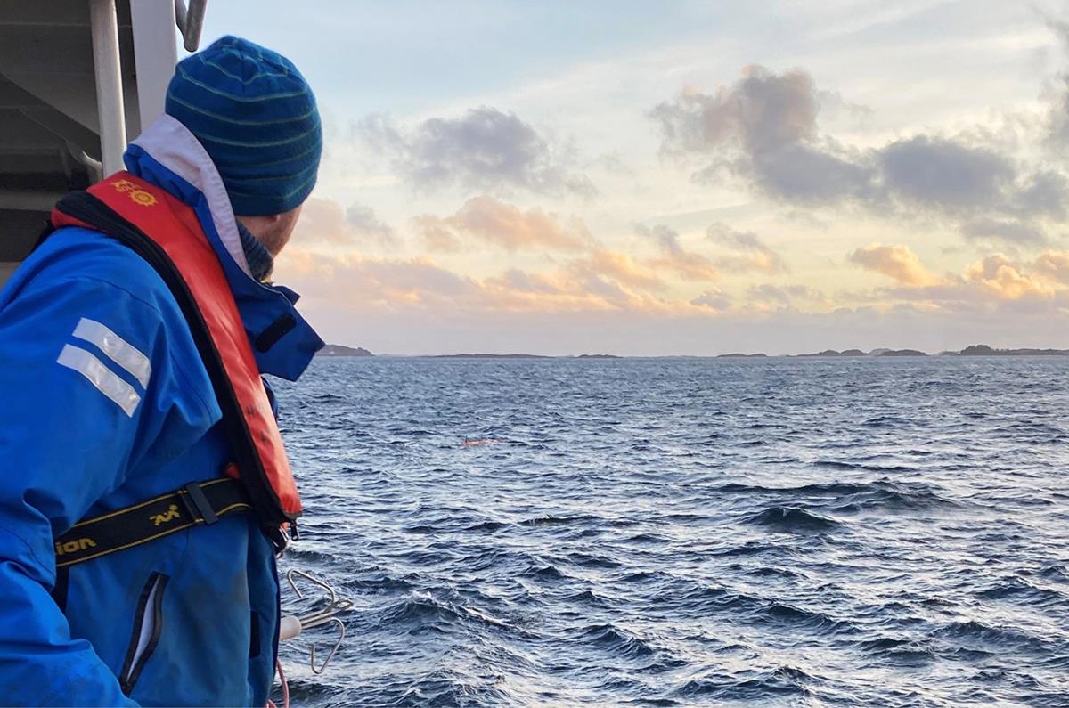 Foto av mannskap på H.U. Sverdrup som speider ut over hav med bølger. Mannen har på seg en blå lue, og rød redningsvest over en blå jakke. I horisonten skimtes en gruppe med øyer. Bildet er tatt i solnedgang og solen er på vei ned bak en sky.