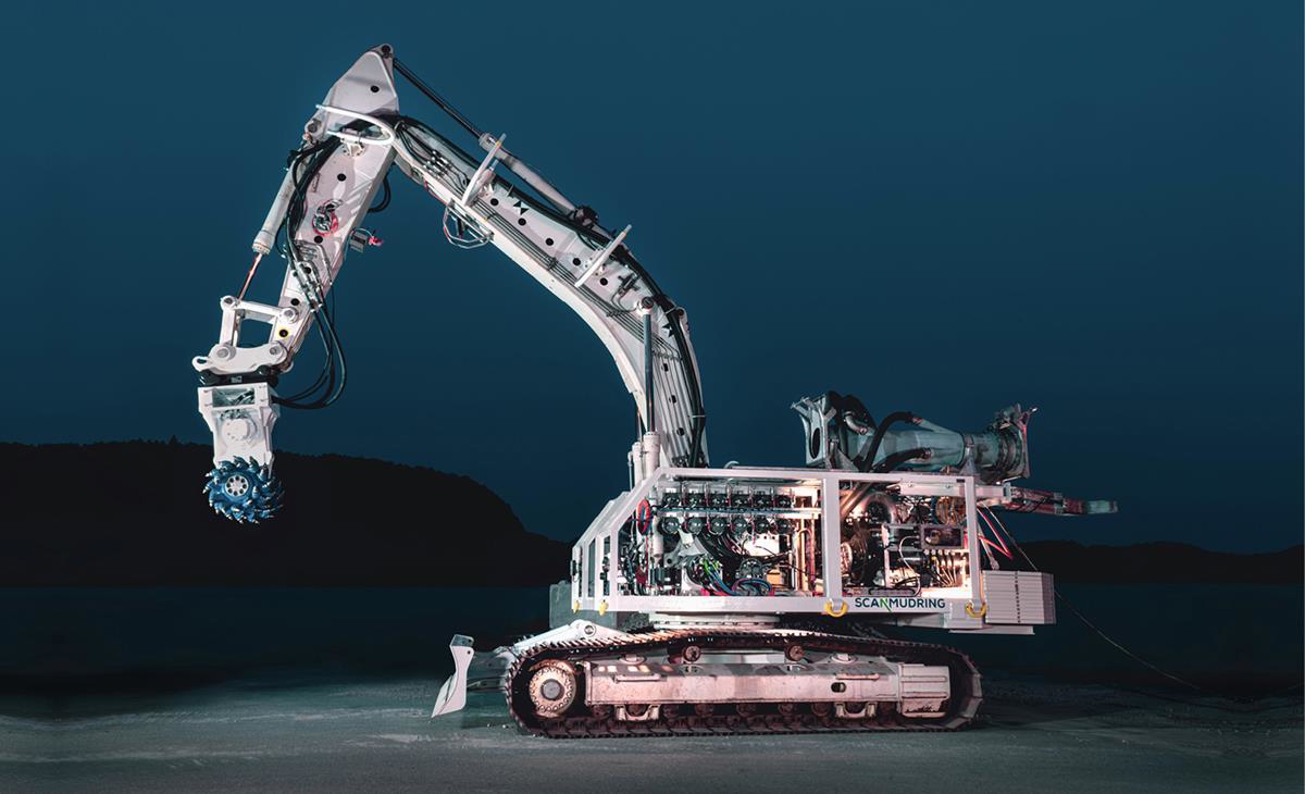 Foto av undervannsgravemaskin. Undervannsgravemaskinen er opplyst, hvit på farge og holder opp grabben. Bildet er tatt om kvelden og bakgrunnen er mørk. Man skimter konturene av land i bakgrunnen.