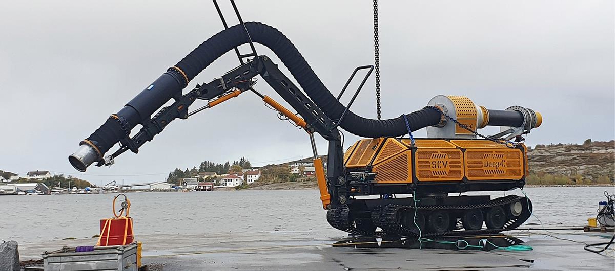 Foto av sugetraktsenhet som står på en kai. Sugetrakten er svart og holdes opp i lufta, mens maskinen er gul. I bakgrunnen ser man grått hav og land med noen hus. Himmelen er grå og overskyet.