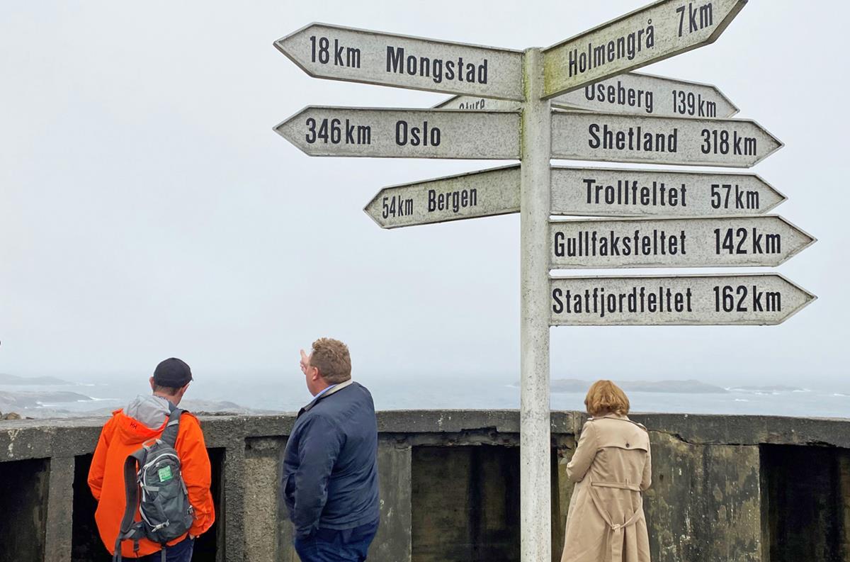 Samlingspunkt på Fedje med skilt som viser avstand til forskjellige kjente oljefelt og steder i Norge. Tre personer ser utover havet. Himmelen er overskyet. I havet ser man flere små holmer.