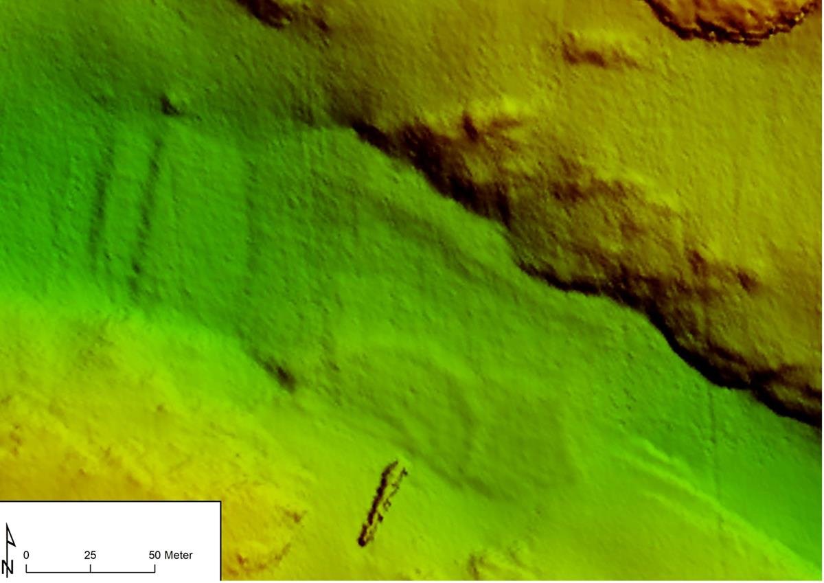 Generert bilde av data fra HUGIN viser havbunnsformasjonen rundt vrakseksjonen illustrert med lysere og mørkere grønnfarger. Støttefyllingen er for det meste vist i den samme grønne fargen, men med stedvis mørkere farger anes konturene på støttefyllingen. Midt på nederst på bildet sees den ene vrakseksjonen. Nede til venstre vises kartets nordpil og målestokk.