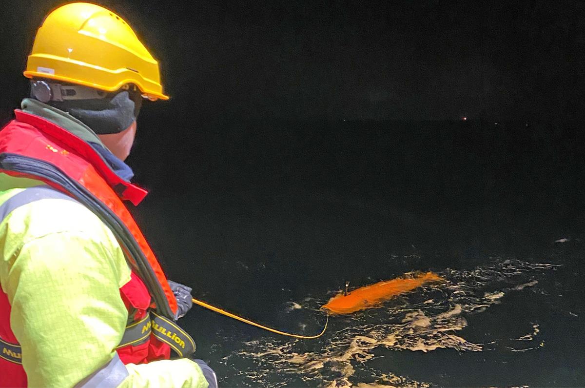 Foto av mannskap på H.U. Sverdrup i ferd med å ta HUGIN opp fra havet. Mannen har på seg hjelm og redningvest, og holder i en gul kabel som er forbundet med HUGIN. Foto er tatt på kvelden og omgivelsene er mørke. Havet har noen skumtopper.