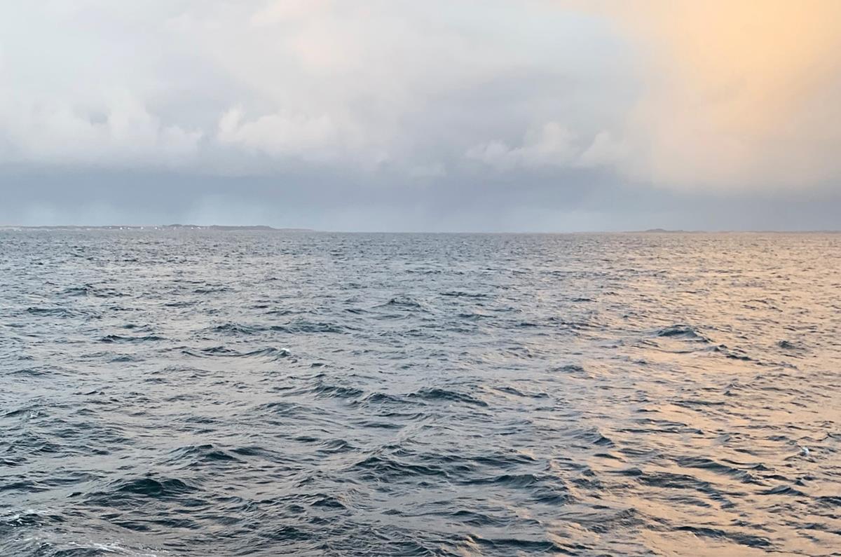 Foto av hav med bølger. I bakgrunnen skimtes en øygruppe. Bildet er tatt ved solnedgang, og himmelen har skyer.