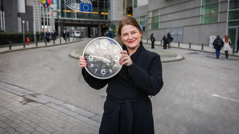 Næringsministeren holder klokke på gata i Brussel med EU-flagg bak.