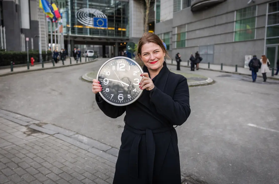 Næringsministeren holder klokke på gata i Brussel med EU-flagg bak.
