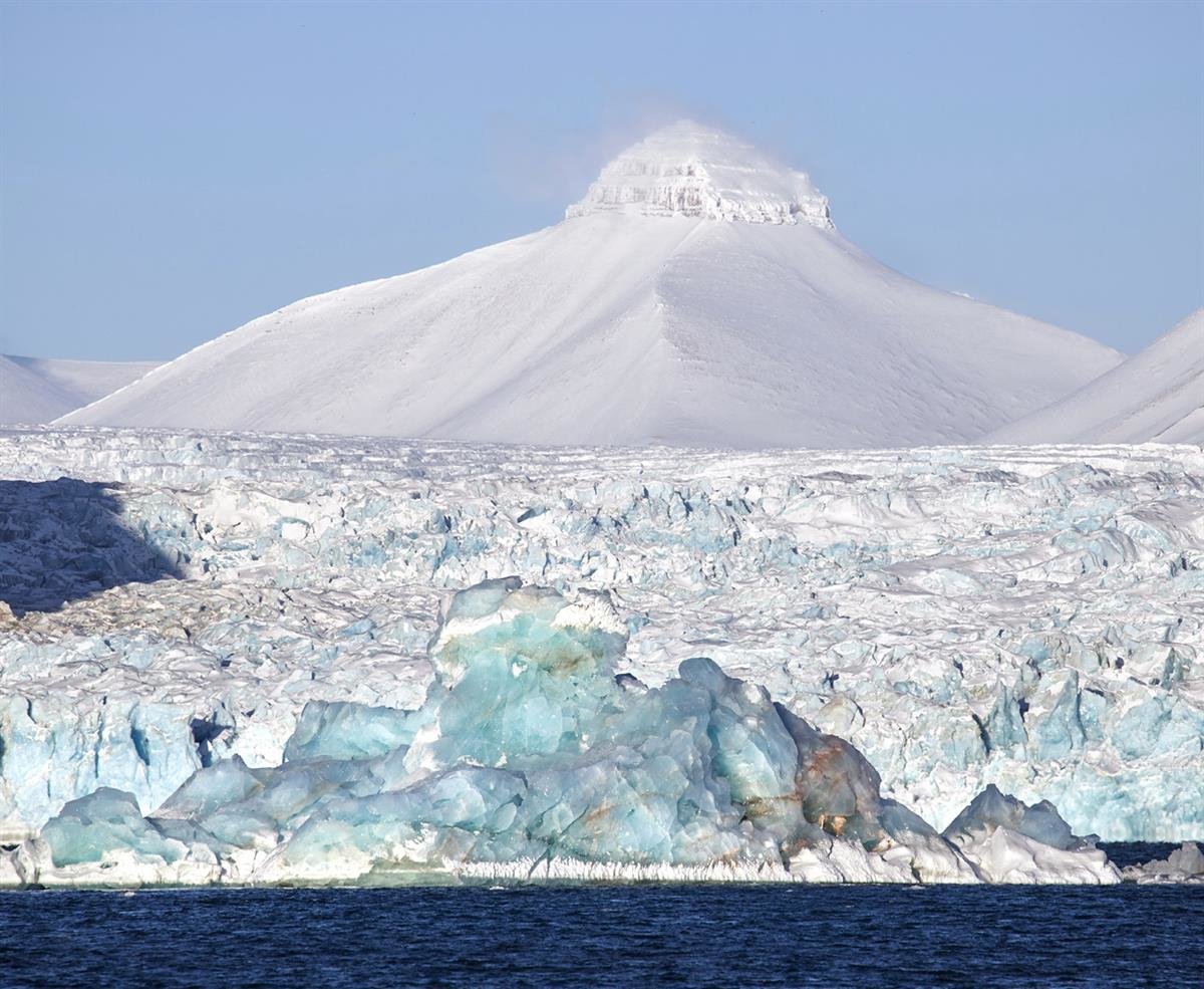 Isfjell i Kongsfjorden og Sveafjellet. Isfjell som flyter foran en isbre. I bakgrunnen ser man fjellet Svea som har en markant liten pyramide av lagdelte bergarter på toppen.  (Utsnitt) Foto: Stein Tronstad / NP