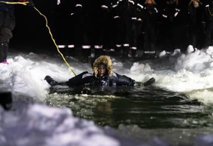 Sikkerhetstrening. En student i beskyttelsesdrakt som ligger i vannet i et stort utgravd hull i isen i mørket, festet i en sikkerhetsline. Flere studenter står i ring rundt og venter på tur. Foto: Ingrid Ballari Nilssen / UNIS