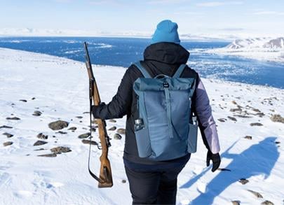Sikkerhet i felt. En person som går i snødekket landskap med fjord og fjell i solskinn med gevær i hånden. Foto: Johanna Schramm / UNIS