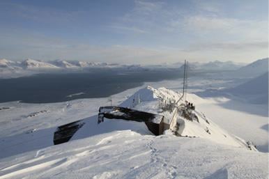 Zeppelinobservatoriet. Luftfoto av et observatoriebygg med antenne ved siden av på toppen av Zeppelinfjellet over Ny-Ålesund, og utsyn over snødekket landskap med fjord og fjell i bakgrunnen. Foto: Max König / NP
