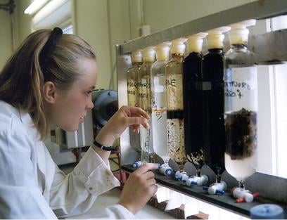 Labarbeid på UNIS. En kvinnelig student i et laboratorium som studerer en rekke glassrør som inneholder vannprøver med forskjellig materiale. To av rørene har svart innhold. Hun måler utenpå det ene røret med en linjal. Foto: UNIS