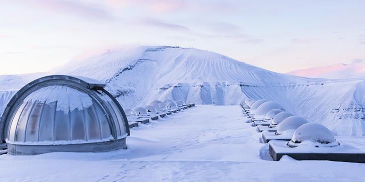 Nordlysobservatoriet ved UNIS. Antenner med runde glasskupler over som står i to rekker på toppen av en bygning på en fjelltopp inne i Adventdalen, i snødekket landskap med fjell i bakgrunnen. Foto: Ingrid Ballari / UNIS